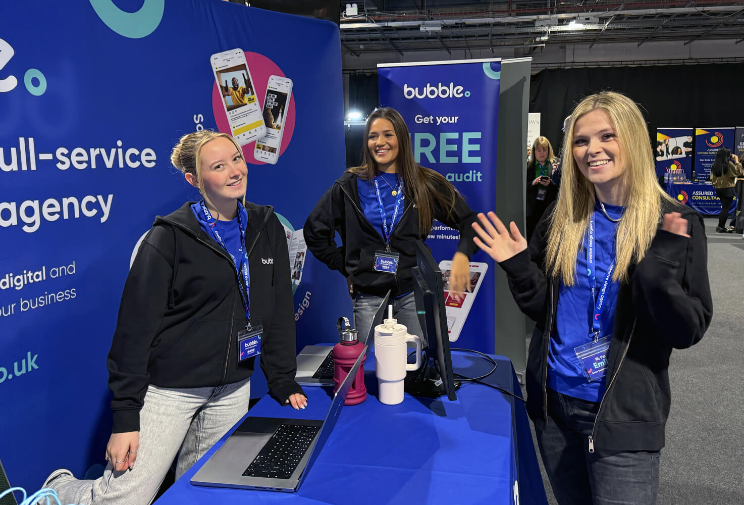 Bubble team members Freya, Izzy and Emily smiling at the camera for a photo in front of the exhibition stand